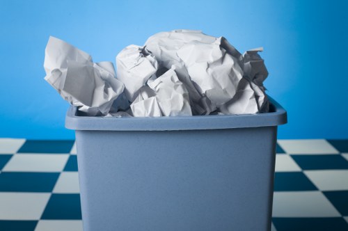 A blue plastic bin filled with crumpled white paper and lightweight waste materials, situated on a checkered black and white tiled floor with a solid blue background. The crumpled paper exhibits various textures and creases, with some pieces extending above the rim of the bin. This scene appears to be indoors, possibly in a room designated for rubbish collection or disposal, and the bin is positioned centrally with no visible surrounding objects. The image emphasizes the process of waste accumulation and disposal relevant to rubbish removal services such as those offered by Garden Clearance Yiewsley, which may operate within the Yiewsley area. The overall lighting is soft, evenly illuminating the contents of the bin and the background, providing a clear and straightforward visual of waste materials prepared for collection or recycling.