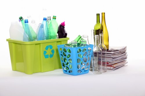 The image features two plastic containers used for recycling or rubbish collection, placed on a white surface. The larger container is lime green with a recycling symbol on its side, filled with various discarded plastic bottles, predominantly in shades of green, blue, and white, with one pink-topped bottle visible. The smaller, bright blue container next to it has a series of circular holes and holds more glass bottles and jars, mostly green and clear, some with remaining labels. To the right of the containers, there is a neat stack of newspapers or magazines with slightly crumpled pages, leaning against two tall, slim glass bottles, one green and one amber, both with narrow necks. The setting appears to be a clean, indoor workspace or a designated rubbish collection area, suitable for a waste management or garden clearance service. This organized display of recyclable materials reflects the context of rubbish removal services offered by Garden Clearance Yiewsley, located near Yiewsley, in the west of London, supporting sustainable waste management practices in the local area.