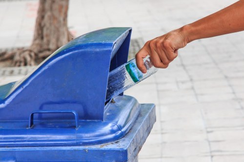 A close-up view of a person's hand discarding a plastic water bottle into a blue communal recycling bin, which is situated outdoors on a paved pavement. The bin features a curved lid that is slightly open, revealing the opening designated for recyclable items. The surrounding environment includes a tree trunk and blurred background that suggests a public space or street scene. The person's hand is visibly grasping the bottle, and the skin tone indicates a fair complexion. The scene captures a moment of waste disposal, consistent with rubbish removal services offered by Garden Clearance Yiewsley in the local area. The lighting is natural, highlighting the textures of the plastic bottle and the smooth surface of the bin, emphasizing environmental practices of recycling and waste separation in the community.