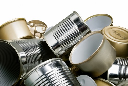 A close-up image displaying a pile of crushed aluminium cans, some with metallic silver finishes and others with gold and white coating, situated on a flat surface likely in an outdoor or industrial setting. The cans are stacked and leaning against each other, with visible ridges and smooth surfaces characteristic of typical household beverage containers. In the background, a grey backdrop suggests a utilitarian environment suitable for rubbish removal activities. The cans show dents and compression marks from being flattened, indicating prior collection or recycling. The lighting is even, highlighting the metallic sheen and reflective qualities of the aluminium, emphasizing their texture and material. This image aligns with the context of waste disposal and recycling services offered by Garden Clearance Yiewsley, particularly for removing scrap metal and beverage cans from domestic or commercial sites around the local Yiewsley area, postcodes nearby.