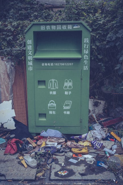 A green, rectangular old clothing donation box positioned outdoors against a backdrop of dark foliage and a wooden fence, with a large pile of discarded shoes, clothing, and miscellaneous rubbish scat