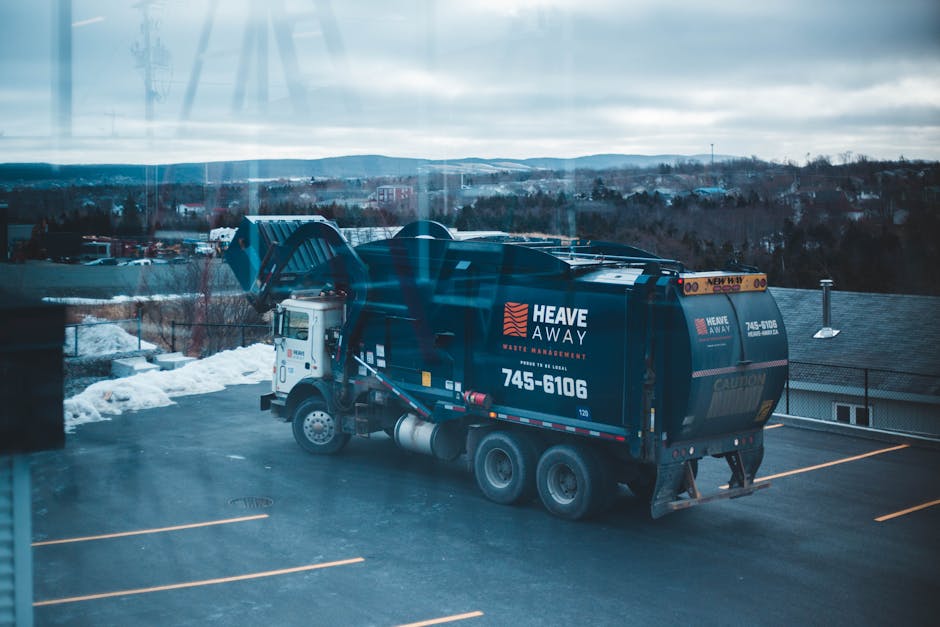 A green, rectangular old clothing donation box positioned outdoors against a backdrop of dark foliage and a wooden fence, with a large pile of discarded shoes, clothing, and miscellaneous rubbish scat